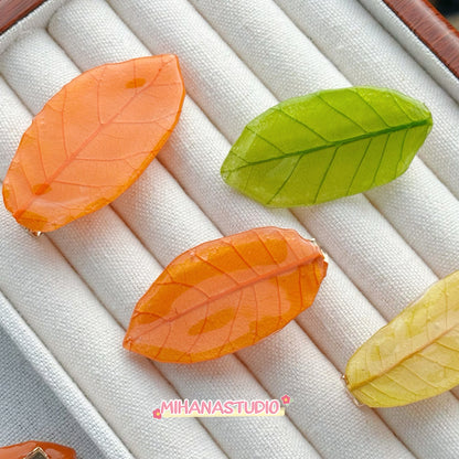 Three leaf-shaped hair clips in orange, green, and yellow on a textured surface with 'MIHANASTUDIO' branding.