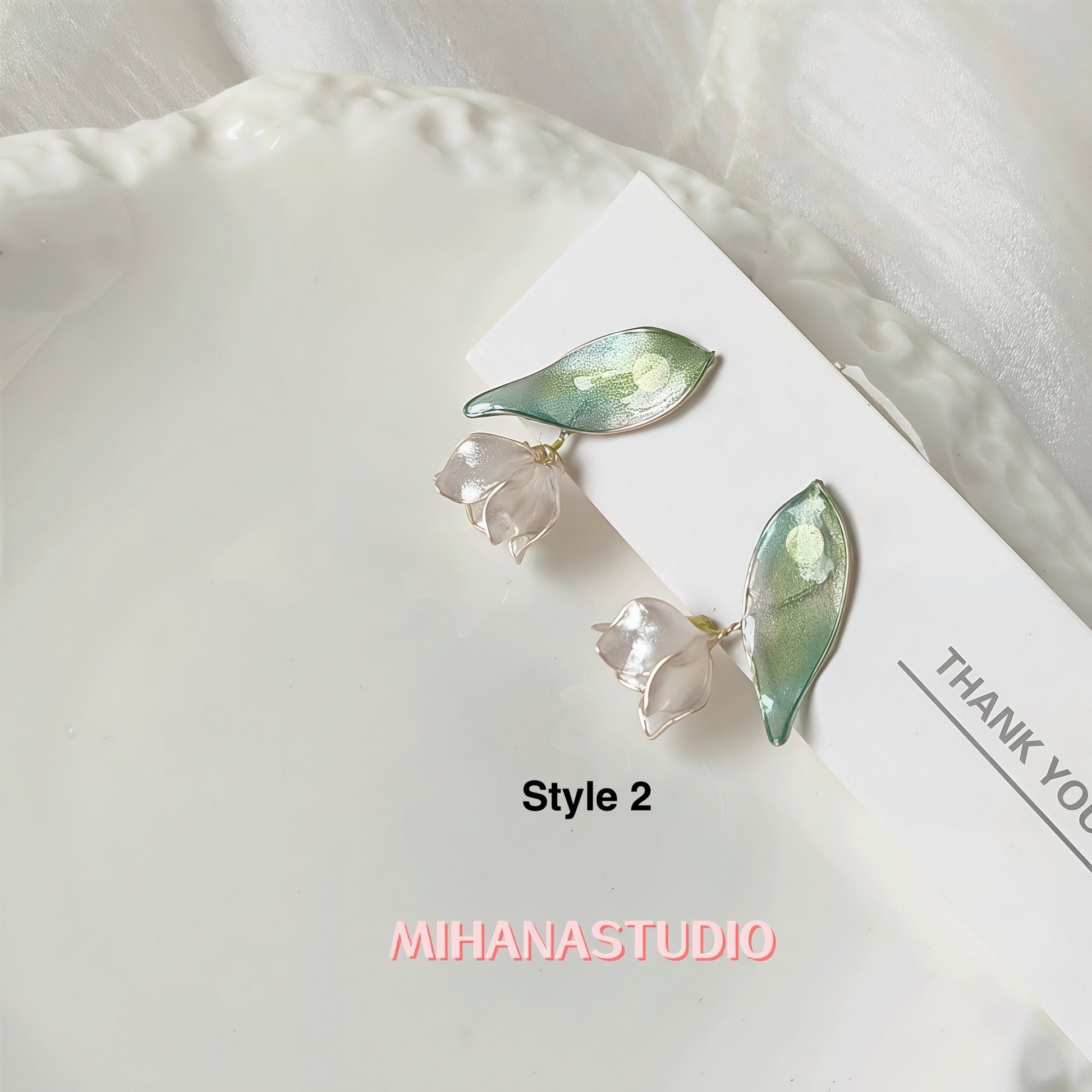 Pair of earrings with leaf and flower design on a white surface, branded 'MIHANASTUDIO'.