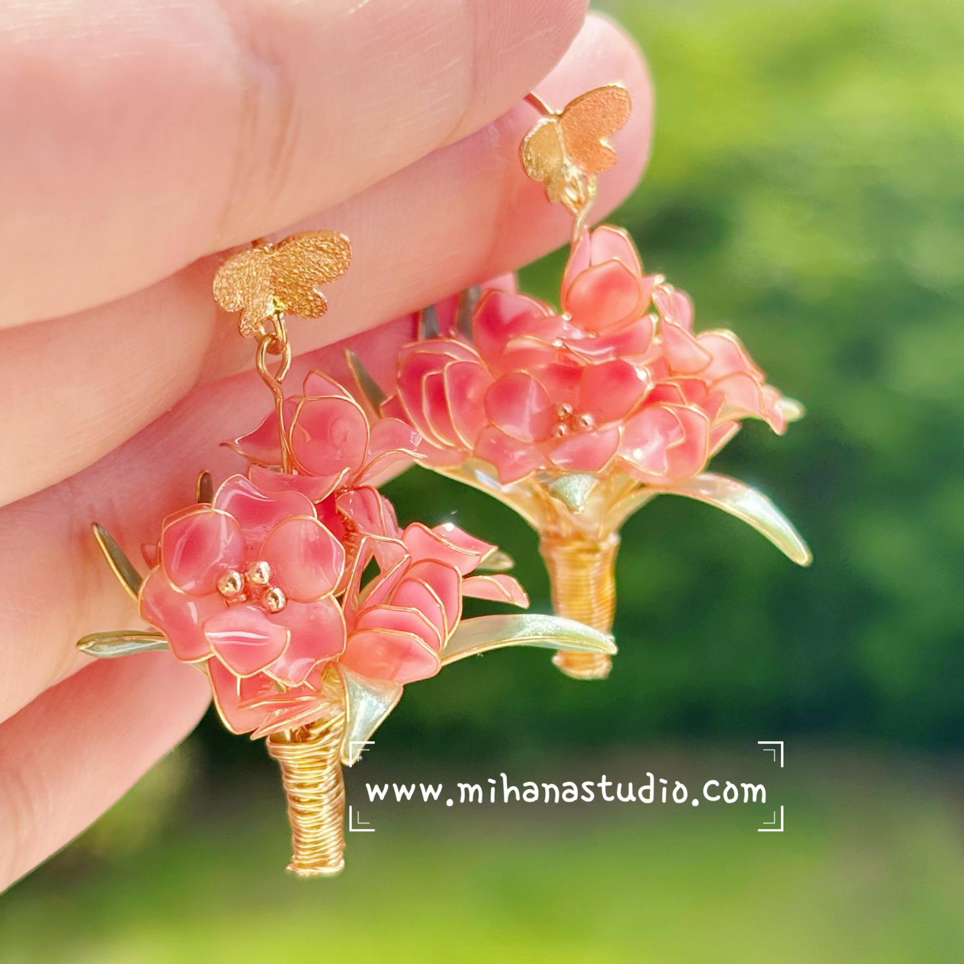 Hand holding a pair of pink floral earrings with gold accents against a blurred green background.