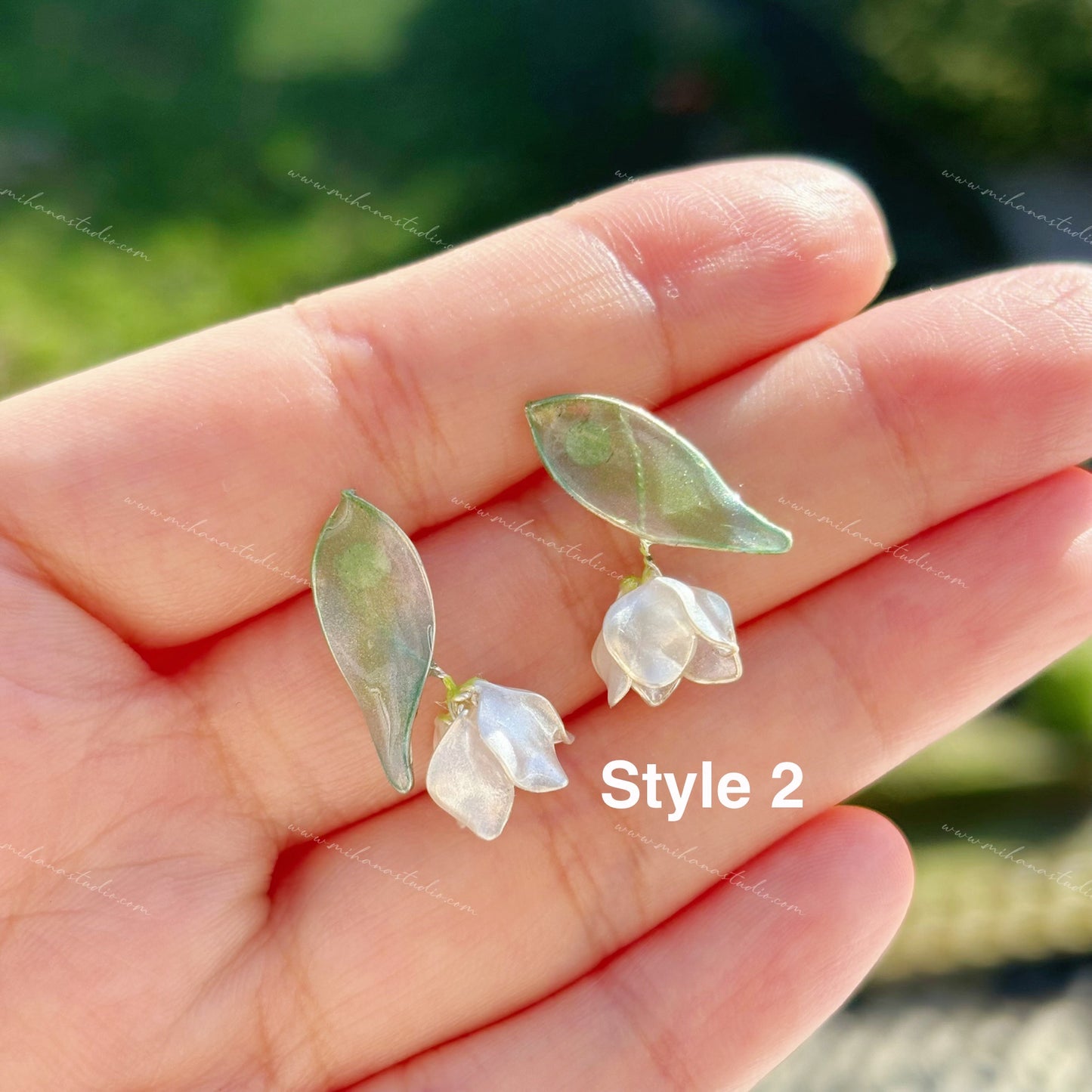 Pair of earrings with green leaves and white flowers held in a hand against a blurred natural background.