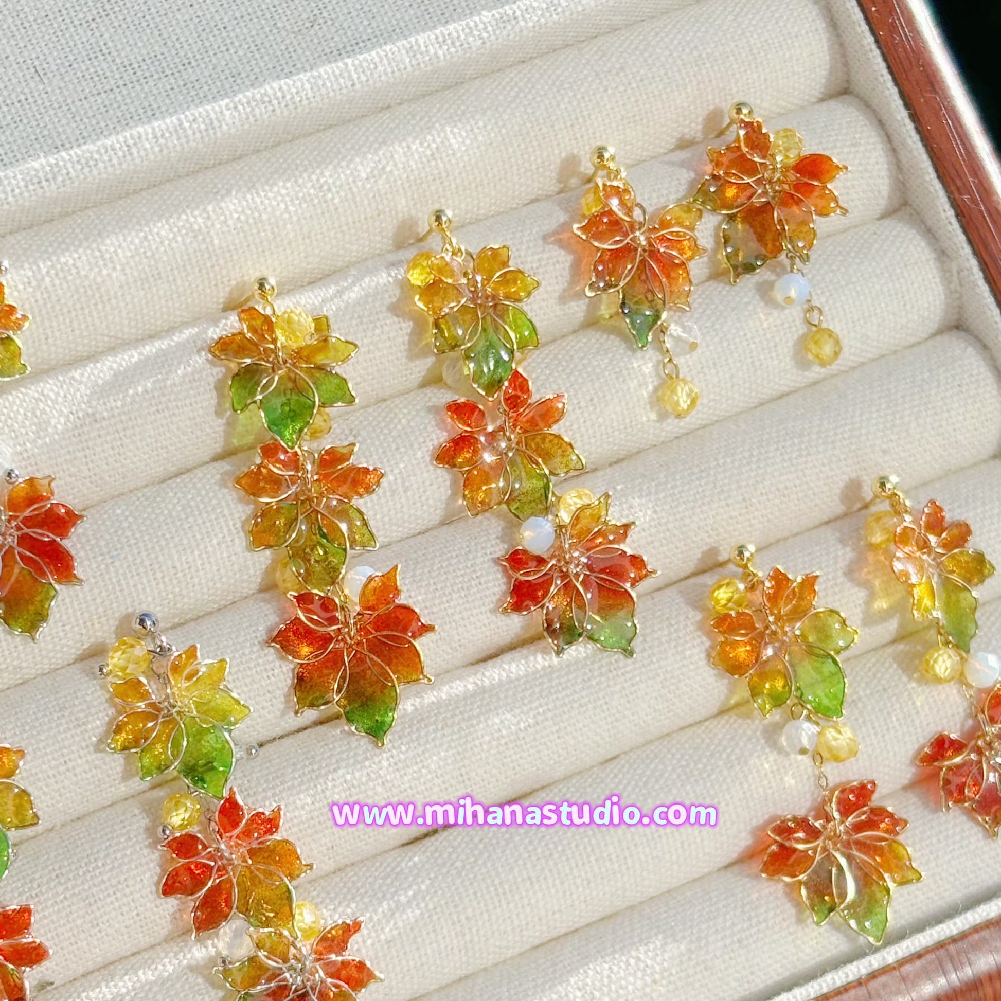 Colourful autumn leaf maple earrings on display in a jewellery box with 'www.mihanastudio.com' branding.