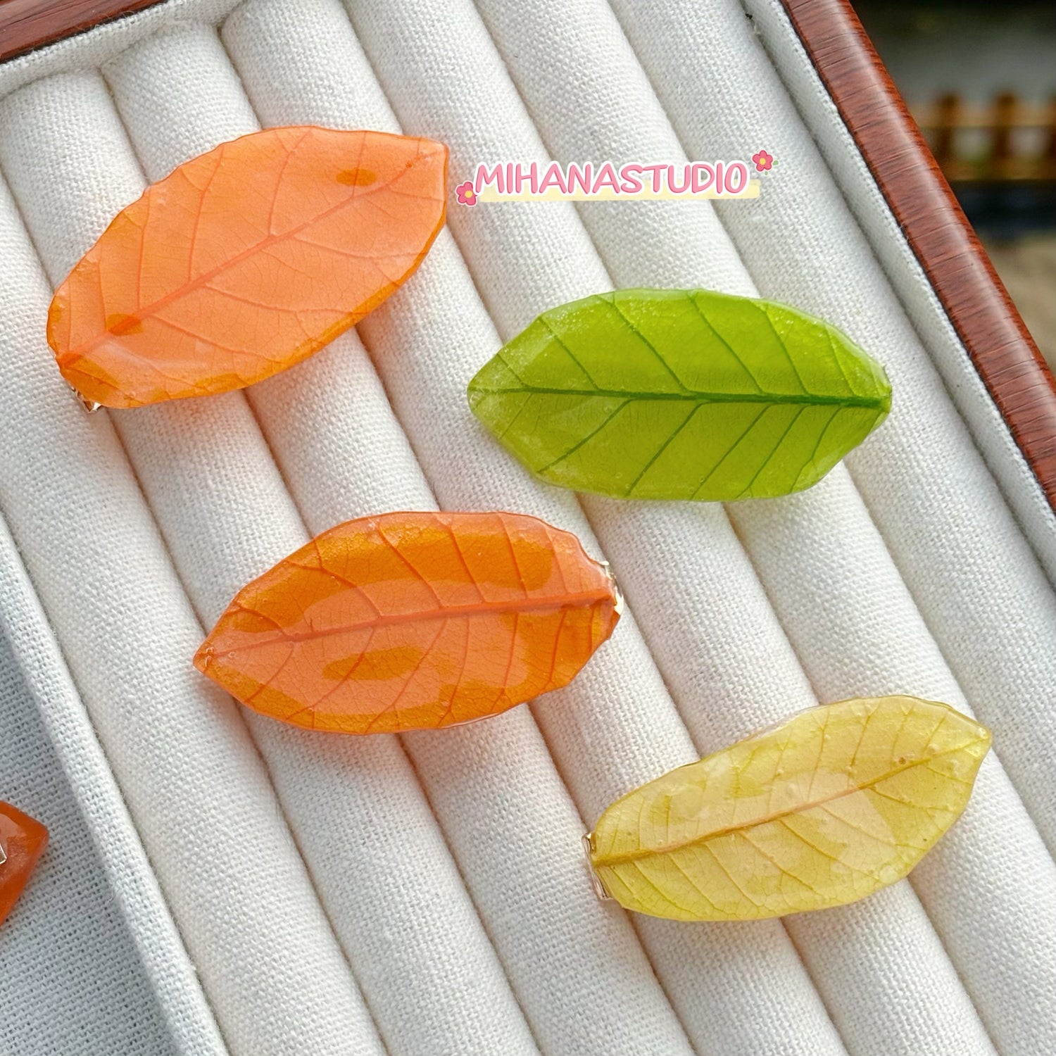 Three leaf-shaped candies in orange, green, and yellow on a textured surface with 'MIHANASTUDIO' branding.