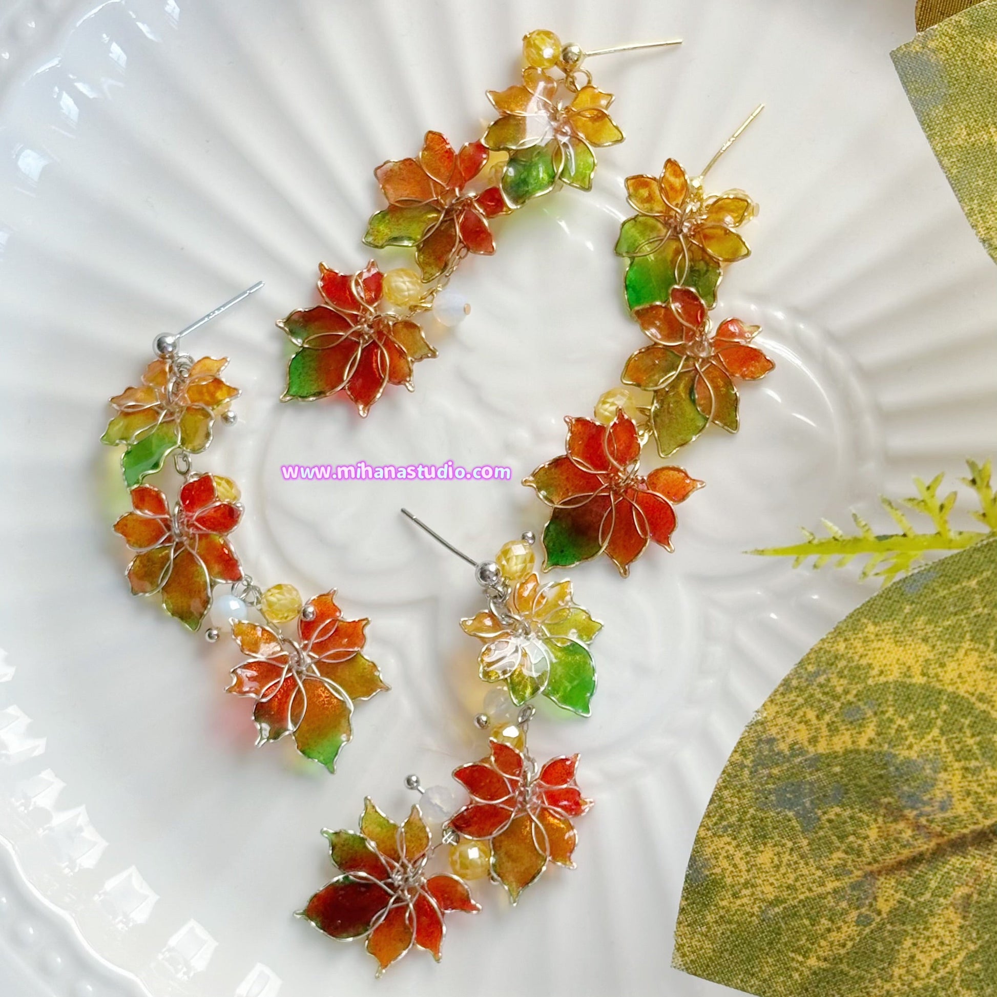 Colourful autumn leaf maple earrings on display in a jewellery box with 'www.mihanastudio.com' branding.