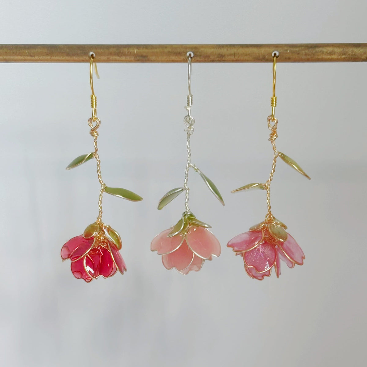 Three dangling flower earrings hang from a wooden rod against a plain light background. Two earrings have pink petals with green leaf-like accents and gold hooks, and the center earring has pale pink petals with a silver hook.