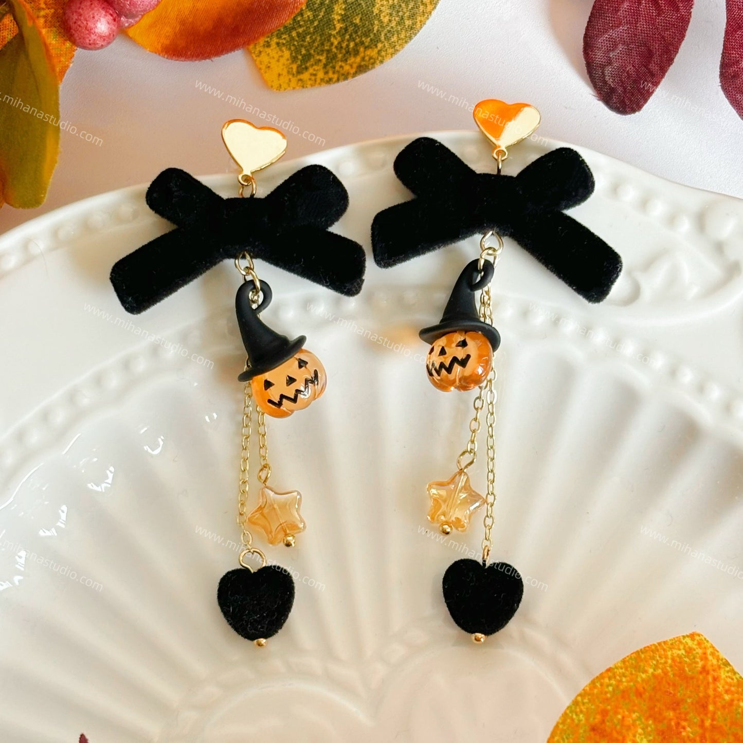 Halloween-themed earrings with black bows, orange pumpkins, and gold hearts on a decorative plate.