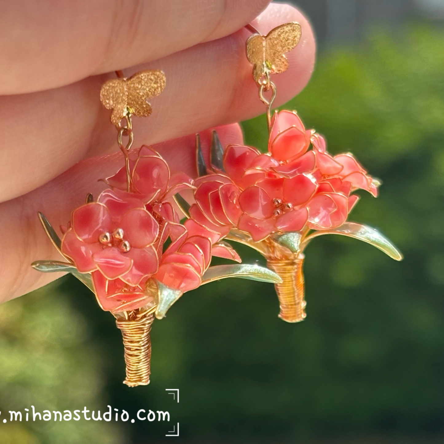 Hand holding a pair of pink floral earrings with gold accents against a blurred green outdoor background.