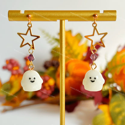 Ghost-shaped earrings with star and bead details on a gold stand against a blurred floral background.