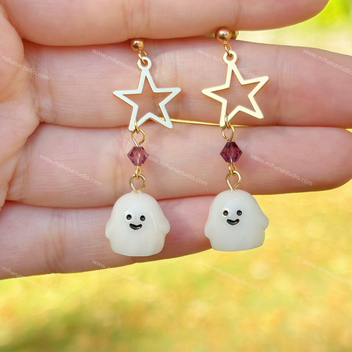 Star-shaped earrings with ghost charms held between fingers against a blurred natural background