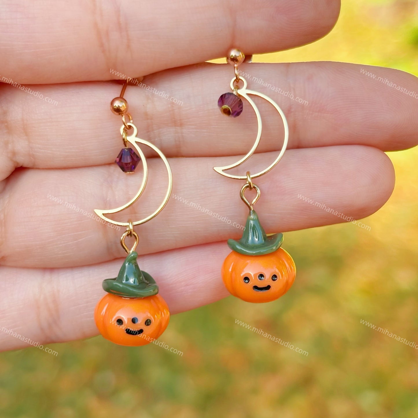 Pumpkin-shaped earrings with green hats held between fingers against a blurred natural background