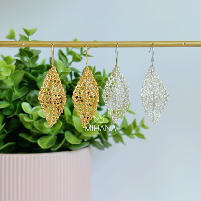 Gold and silver earrings hanging on a rod with green plants in the background, featuring the brand 'MIHANA'.