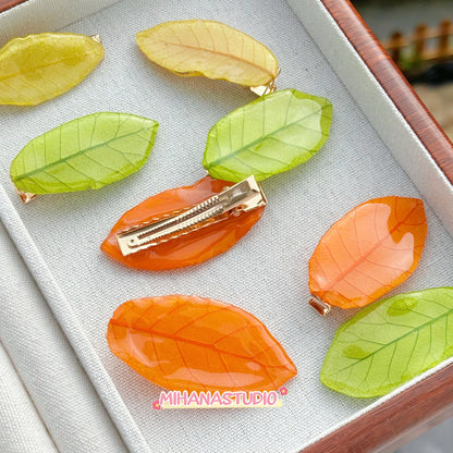 Leaf-shaped hair clips in orange and green on a textured surface with 'MIHANA STUDIO' branding.