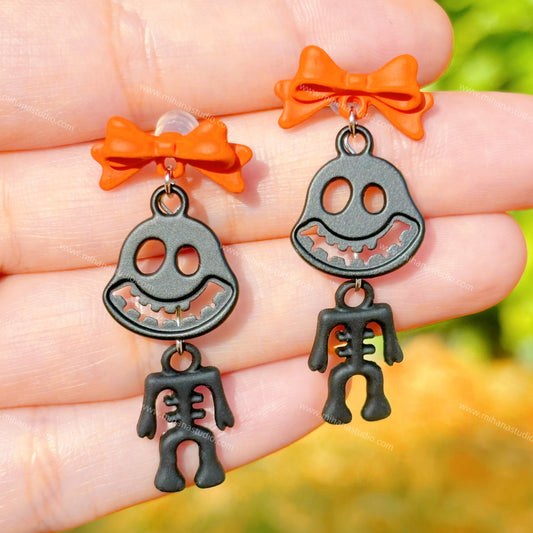 Halloween-themed earrings with skeleton design and orange bows held in a hand against a blurred natural background.