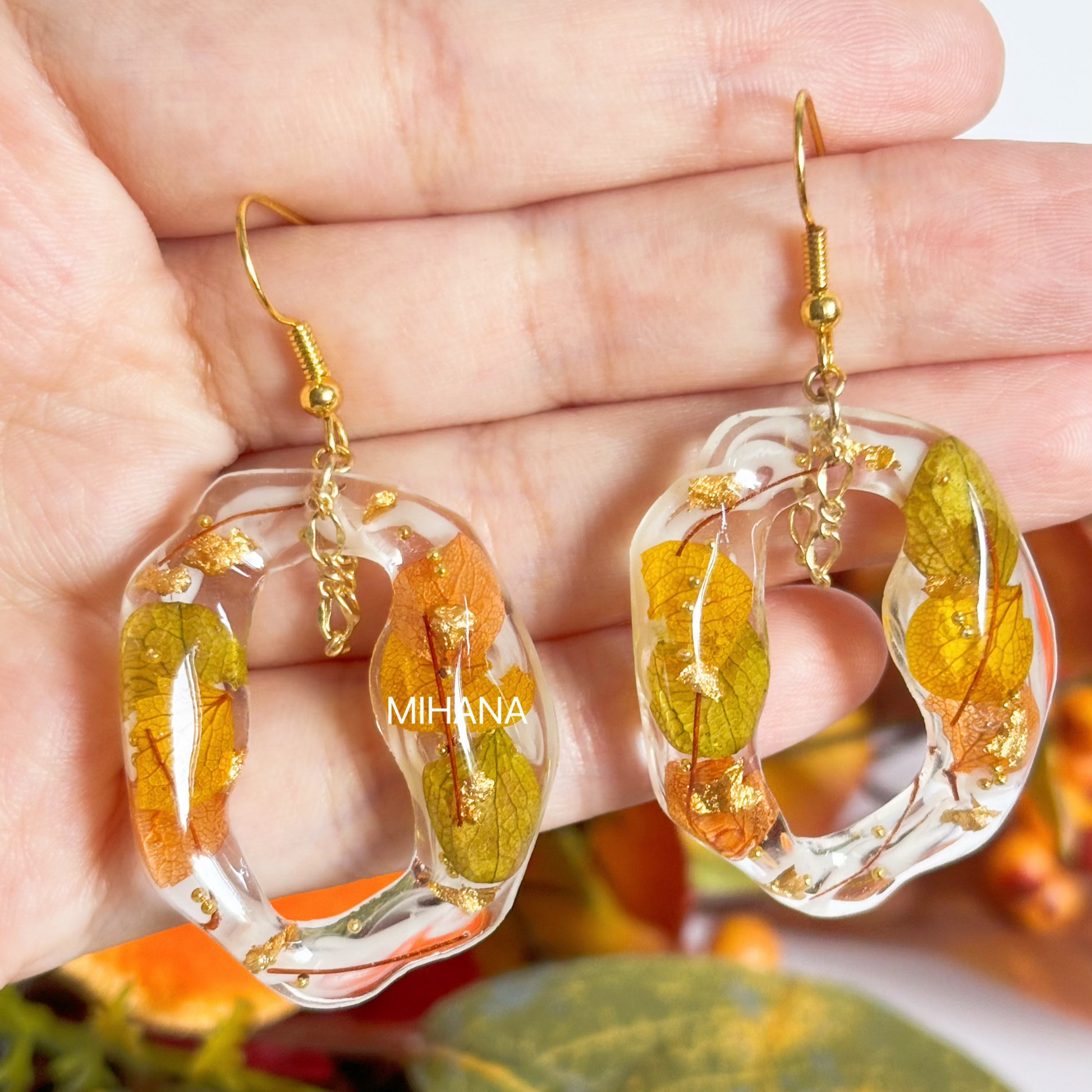 Clear hoop earrings with leaf design held between fingers, brand 'MIHRA' visible.