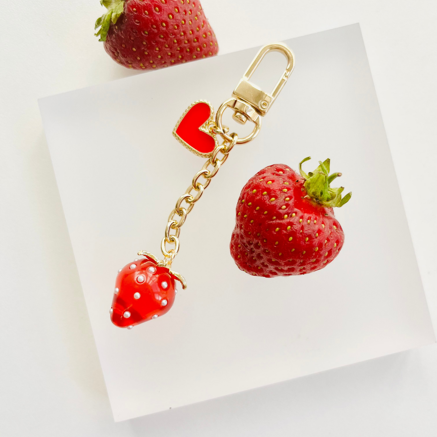 A red resin strawberry bagcharm attached to a gold-colored keychain, displayed next to a real strawberry on a white surface.