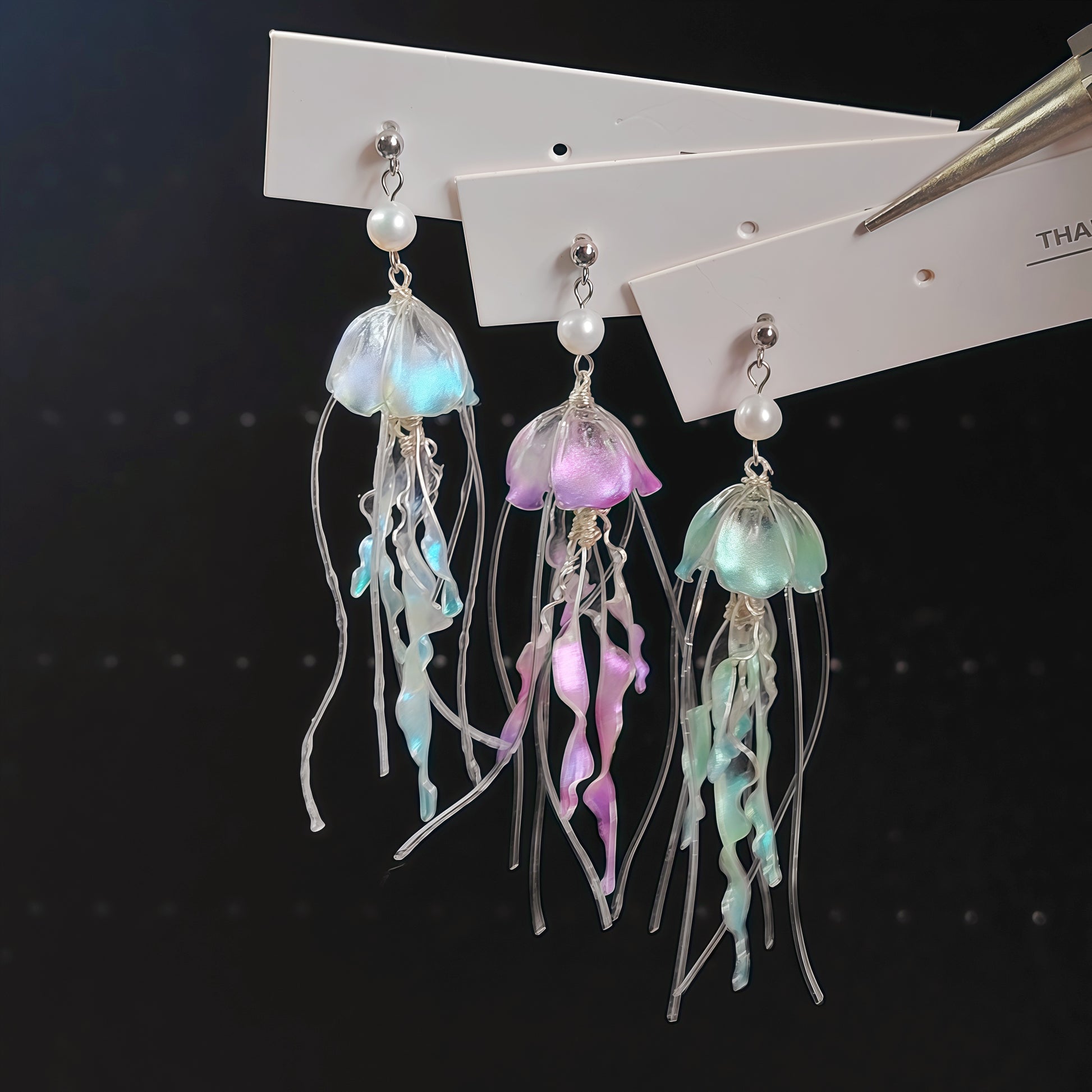 Three jellyfish-shaped dangling earrings displayed on white cards against a dark background. The earrings have iridescent translucent bell shapes in pastel blue, purple, and mint green, with long flowing tentacles and small white pearl-like beads above silver hooks.