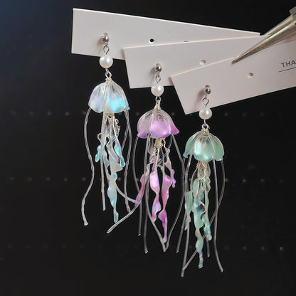 Three jellyfish-shaped dangling earrings displayed on white cards against a dark background. The earrings have iridescent translucent bell shapes in pastel blue, purple, and mint green, with long flowing tentacles and small white pearl-like beads above silver hooks.